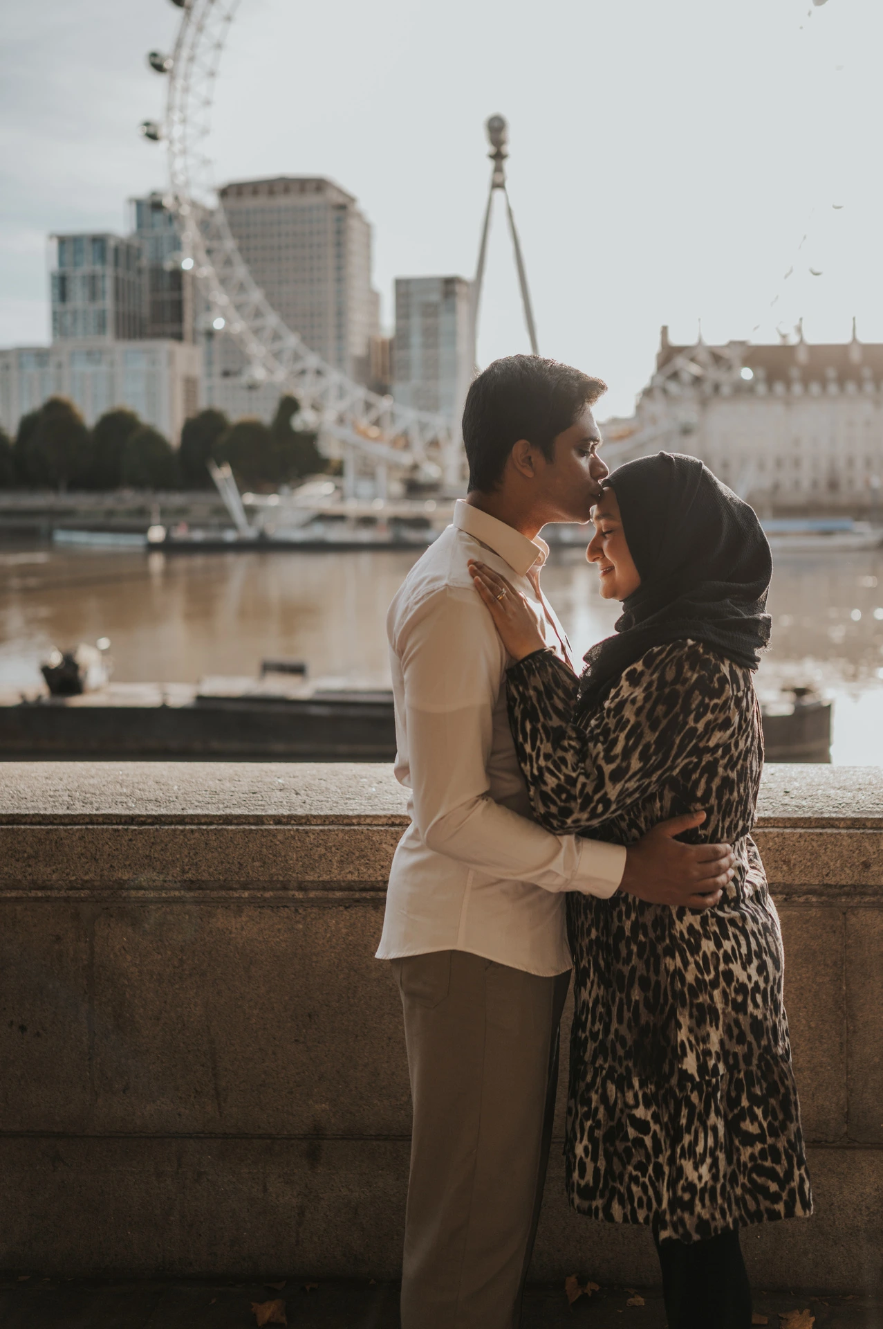 Couple standing in an embrace beside the Thames at Westminster Pier, with the London Eye behind them, during their London photoshoot.