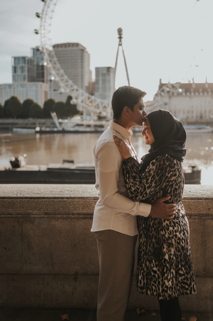 London couple photoshoot embracing by the Thames Couple standing in an embrace beside the Thames at Westminster Pier, with the London Eye behind them, during their London photoshoot.