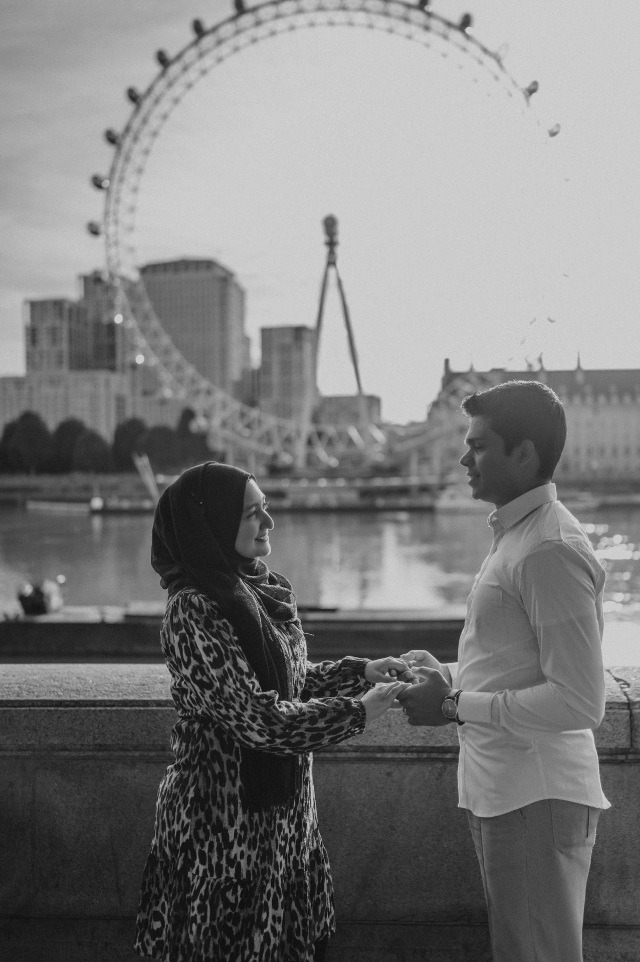 Black-and-white portrait of the couple holding hands by the stone wall at Westminster Pier, with the London Eye behind them, from their London photoshoot.