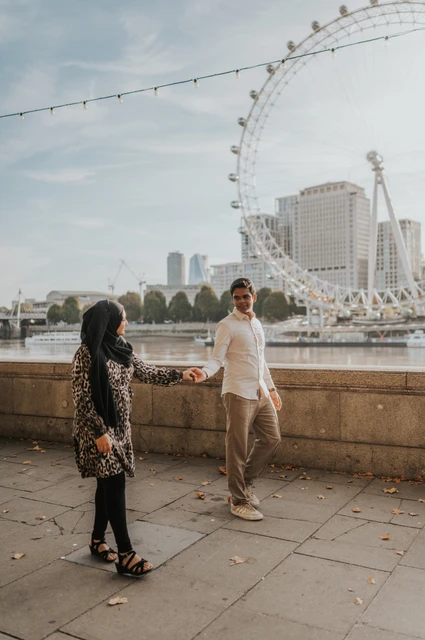 London couple photoshoot holding hands by Westminster Pier Full-length photograph of the couple holding hands beside the Thames at Westminster Pier, with the London Eye behind them, during their London photoshoot.