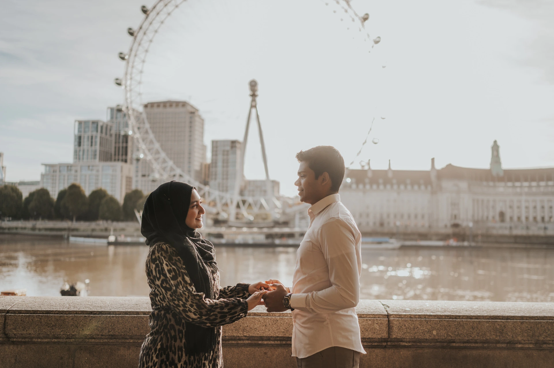 Couple holding hands beside the sunlit Thames at Westminster Pier, with the London Eye behind them, during their London photoshoot.
