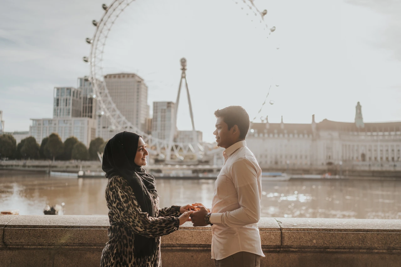 London couple photoshoot in warm riverside light Couple holding hands beside the sunlit Thames at Westminster Pier, with the London Eye behind them, during their London photoshoot.