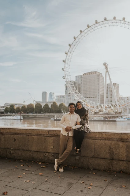 Full-length portrait of the couple together beside the stone riverside wall at Westminster Pier, with the London Eye across the Thames, during their London photoshoot.