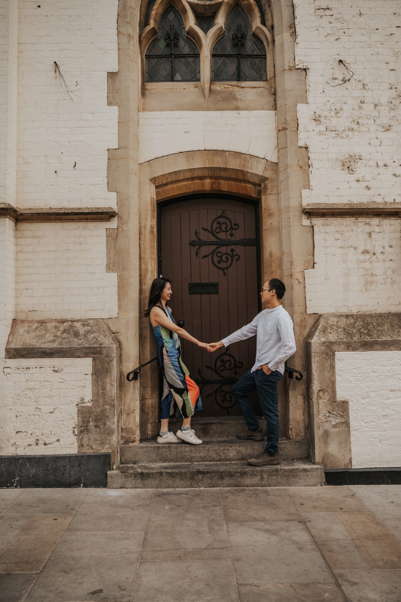 The couple hold hands across the stone steps of an arched wooden doorway framed by pale stonework during their London couple photoshoot.