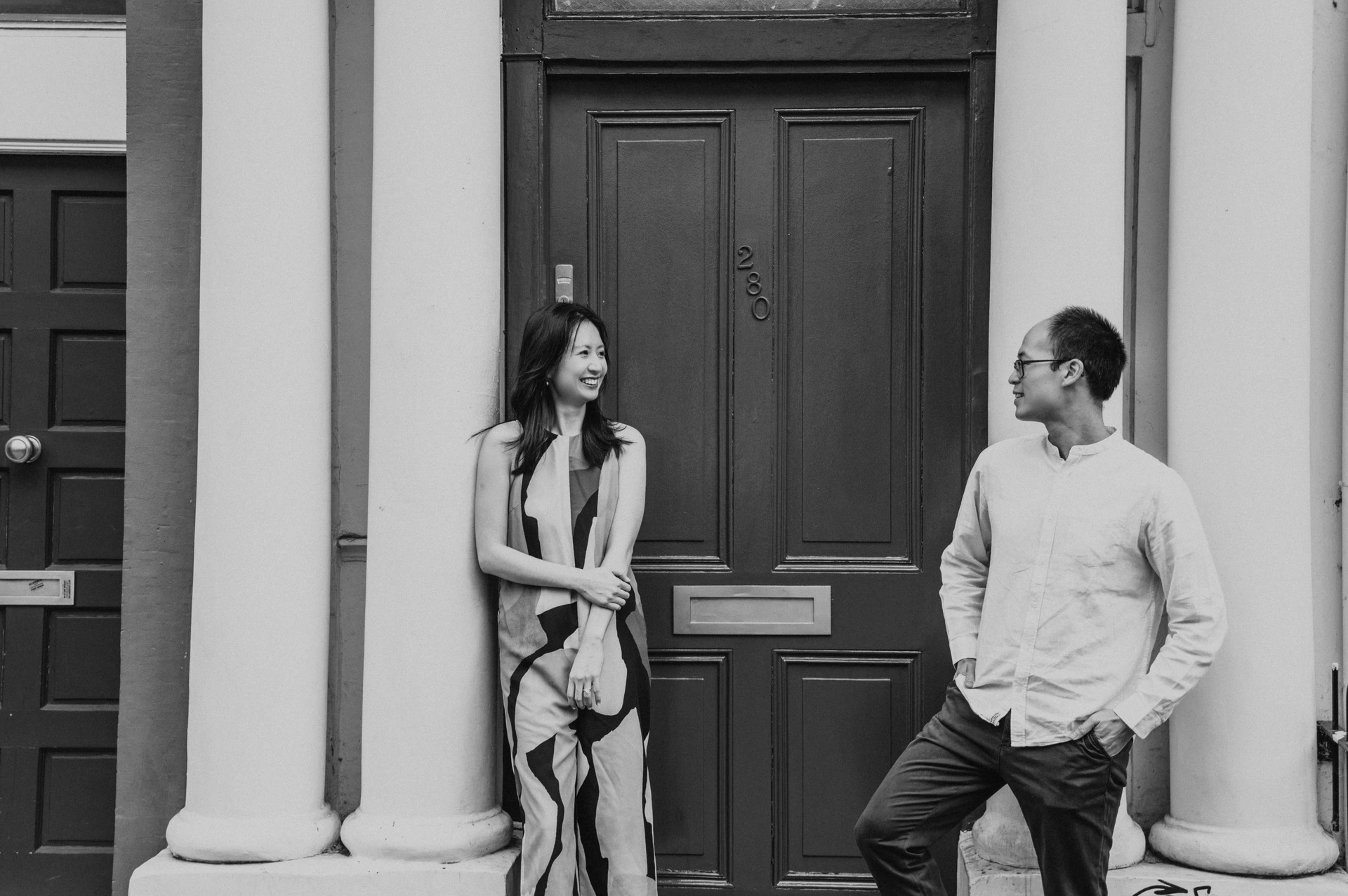 A black-and-white wide portrait of the couple standing on either side of a front door and looking at each other during their London couple photoshoot.