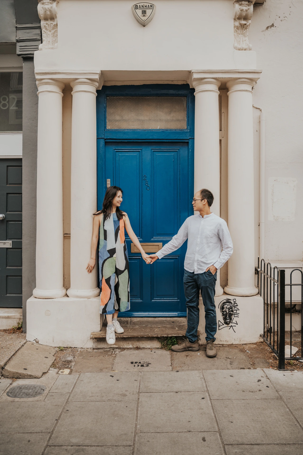 The couple stand on either side of a bright blue front door and hold hands beneath its cream stone columns during their London couple photoshoot.