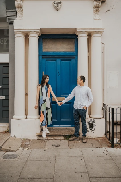 London couple photoshoot: holding hands beneath the blue doorway columns The couple stand on either side of a bright blue front door and hold hands beneath its cream stone columns during their London couple photoshoot.