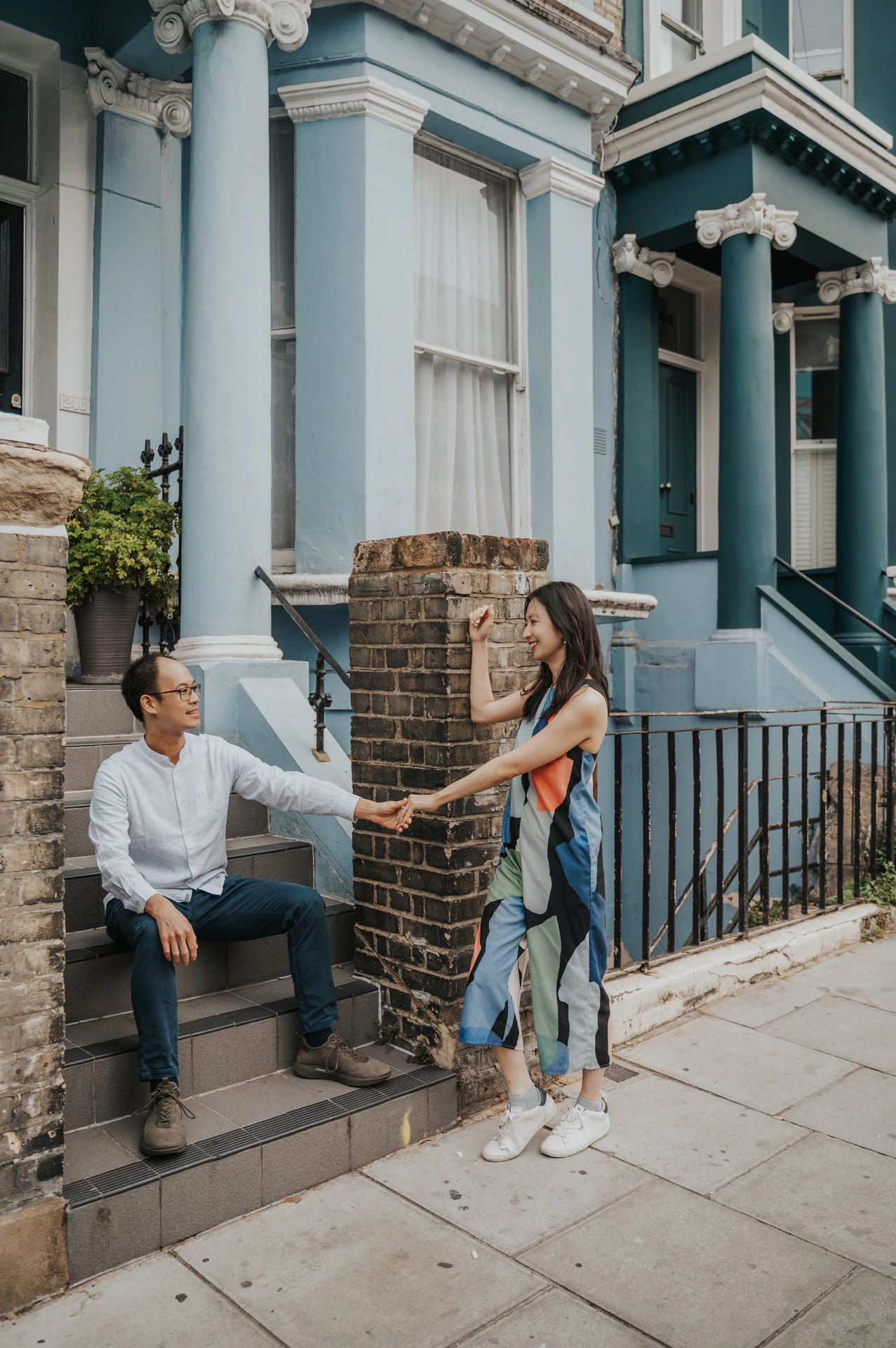 He sits on stone steps while she stands holding his hand beside the blue and green Notting Hill houses during their London couple photoshoot.