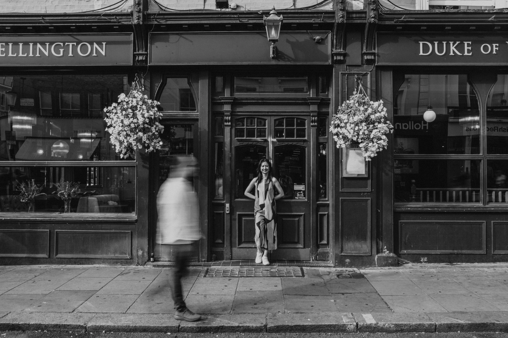 A black-and-white view of the couple outside the Duke of Wellington pub with a blurred pedestrian passing in the foreground during their London couple photoshoot.