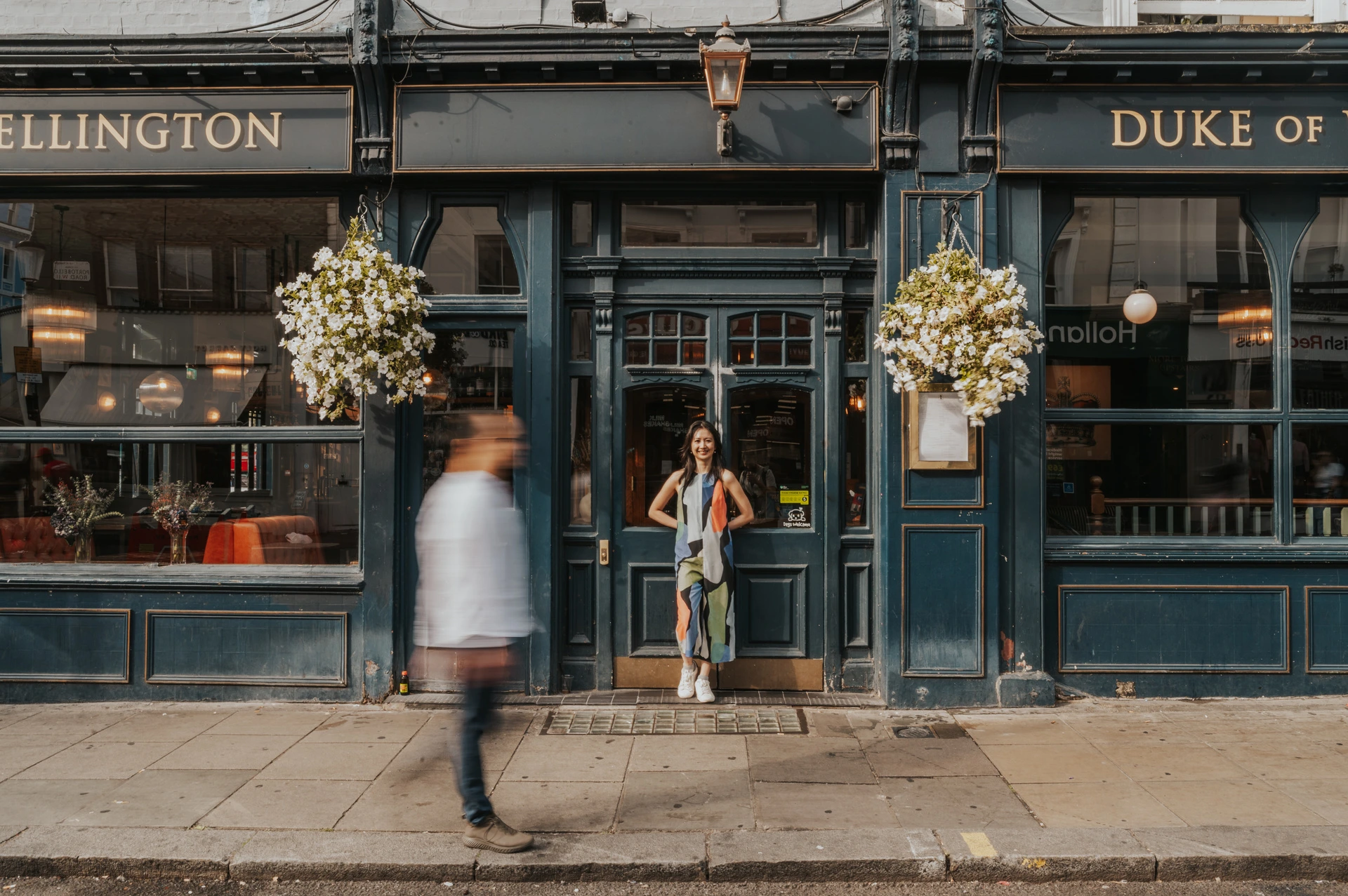 The couple stand close together outside the dark green Duke of Wellington pub while a blurred pedestrian crosses the foreground during their London couple photoshoot.