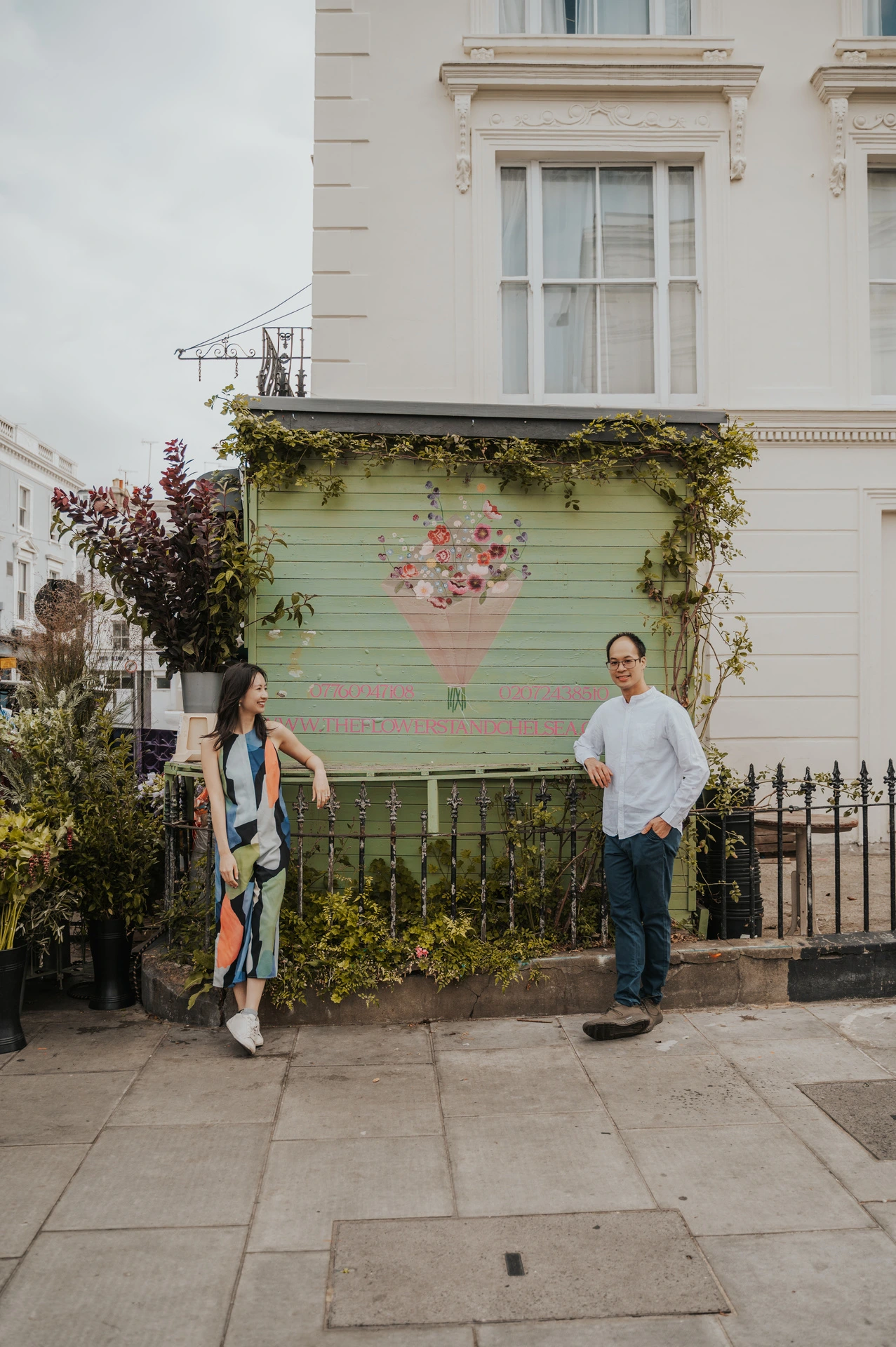 The couple stand apart and smile beside a green shop shutter painted with a bouquet of flowers during their London couple photoshoot.