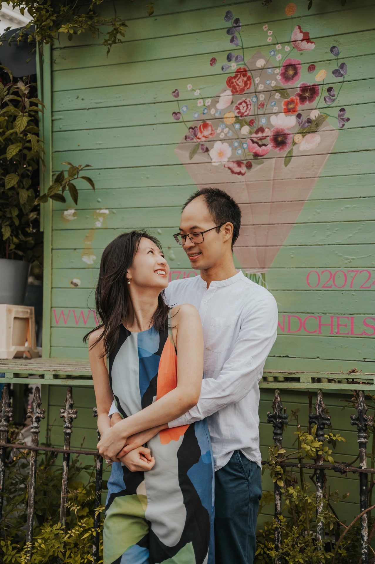 He embraces her from behind as she looks up at him in front of a green shutter painted with flowers during their London couple photoshoot.