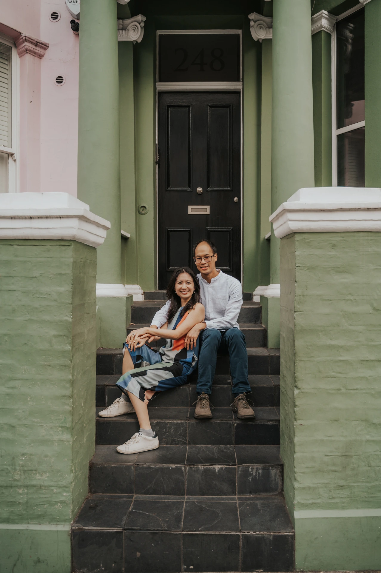 The couple sit together on the stone staircase beneath the black front door of a green Notting Hill house during their London couple photoshoot.