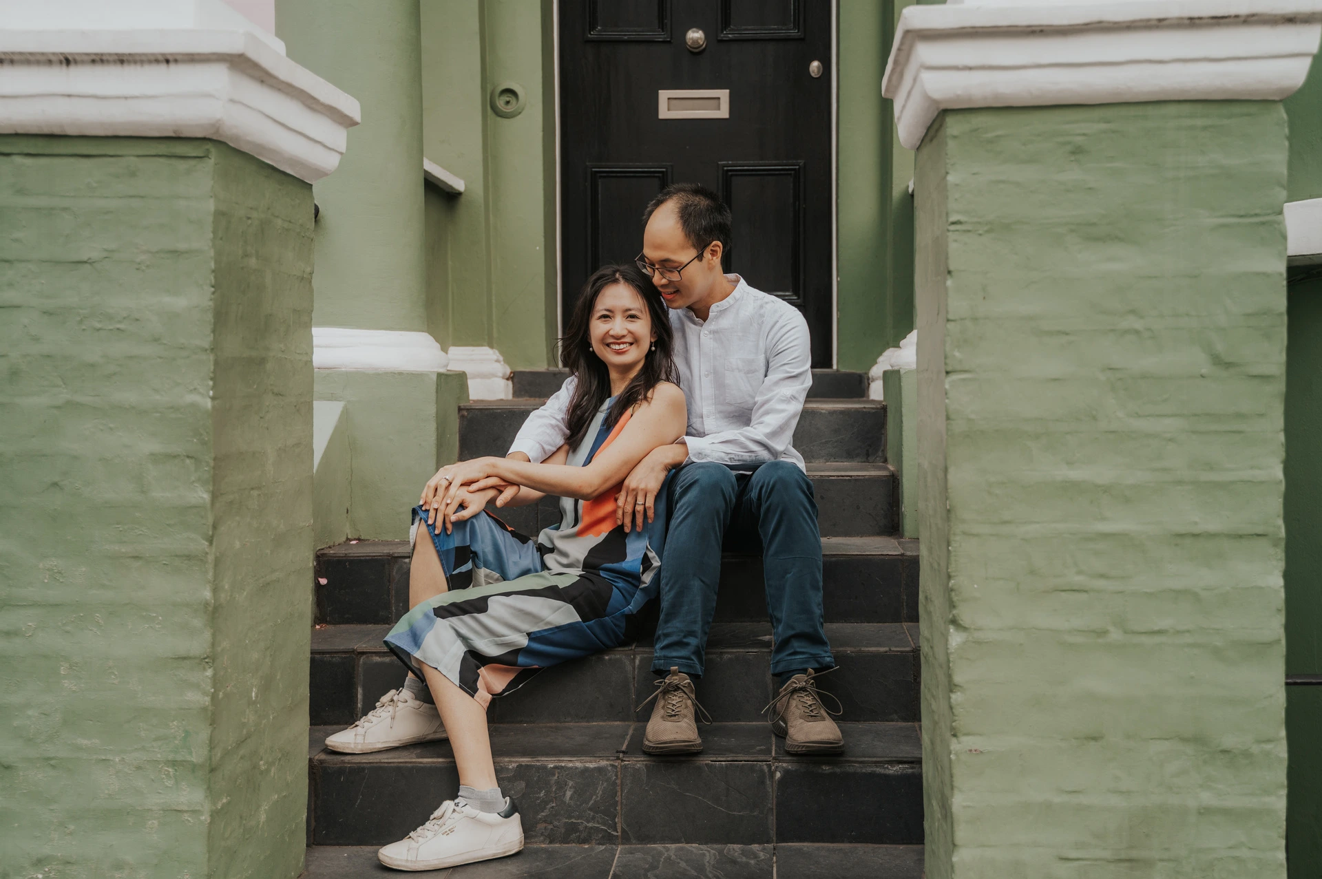 A wide view of the couple sitting together on the steps between the green house walls and brick gateposts during their London couple photoshoot.