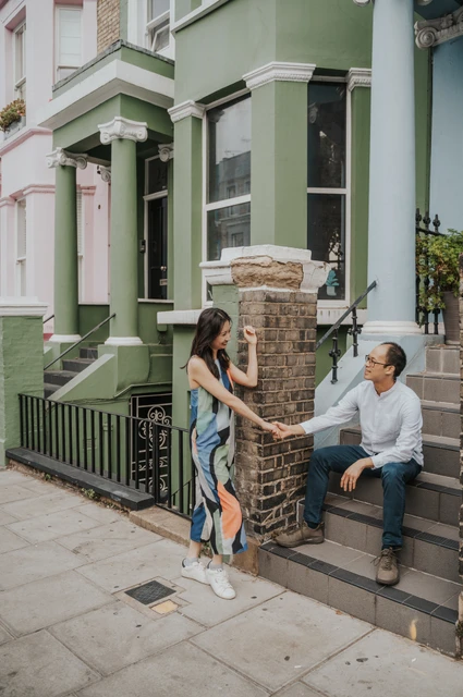 London couple photoshoot: holding hands by the green house steps She stands beside a brick gatepost and holds his hand while he sits on the steps of a green Notting Hill house during their London couple photoshoot.
