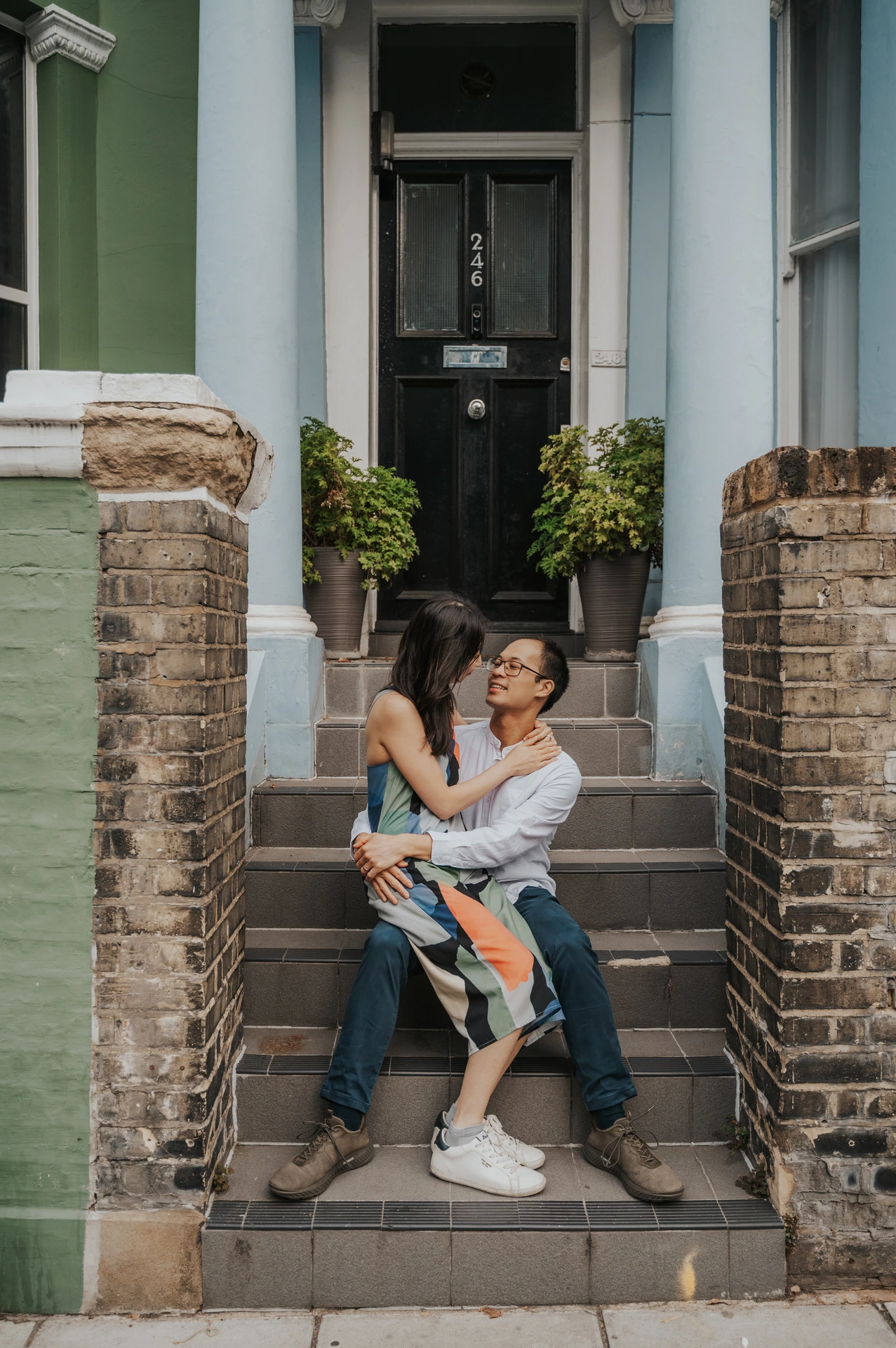 She sits on his lap on the stone steps of a green Notting Hill house, and they smile towards each other during their London couple photoshoot.