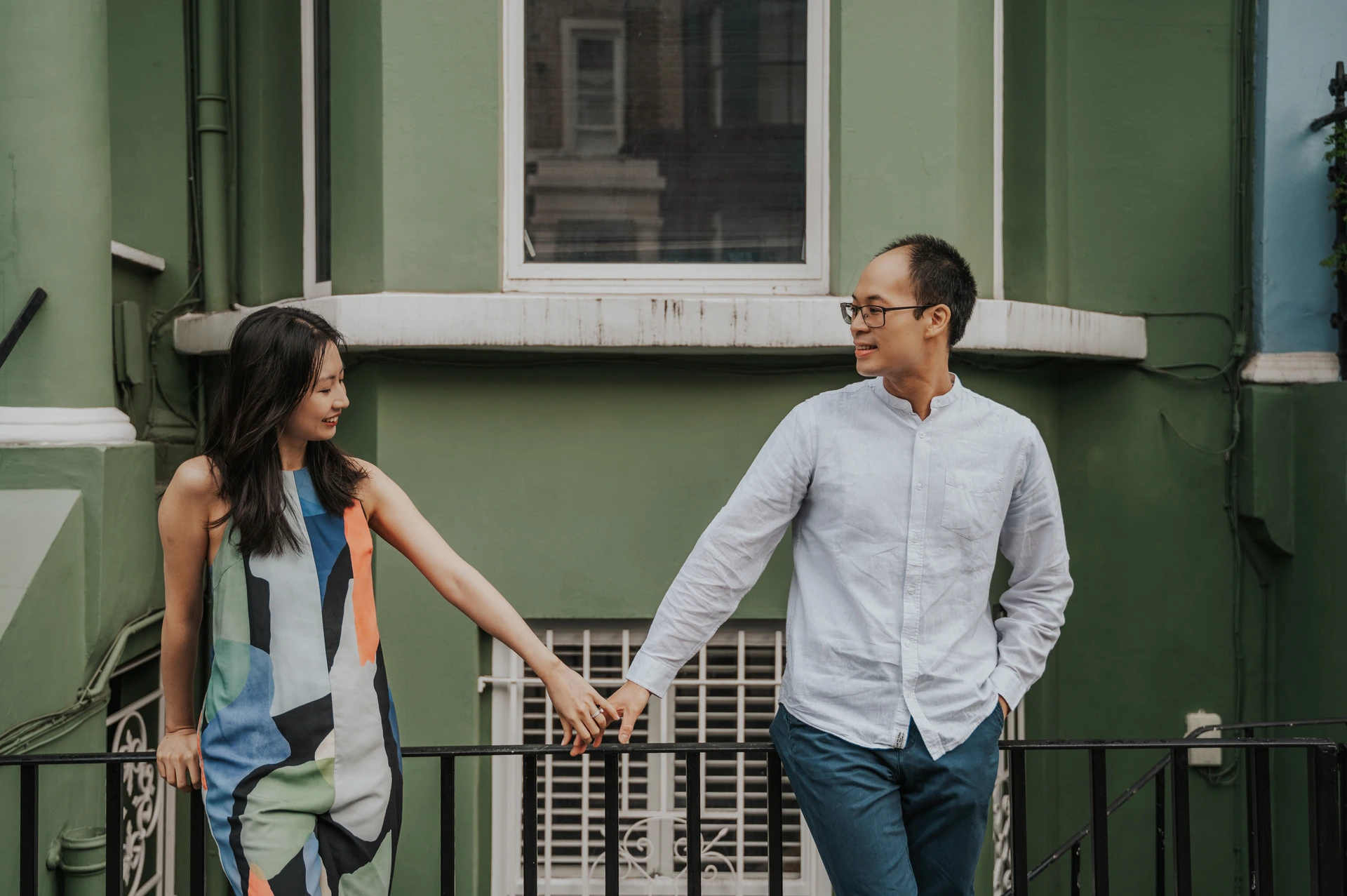 A wide portrait of the couple holding hands and looking at each other in front of the green wall and black railings during their London couple photoshoot.