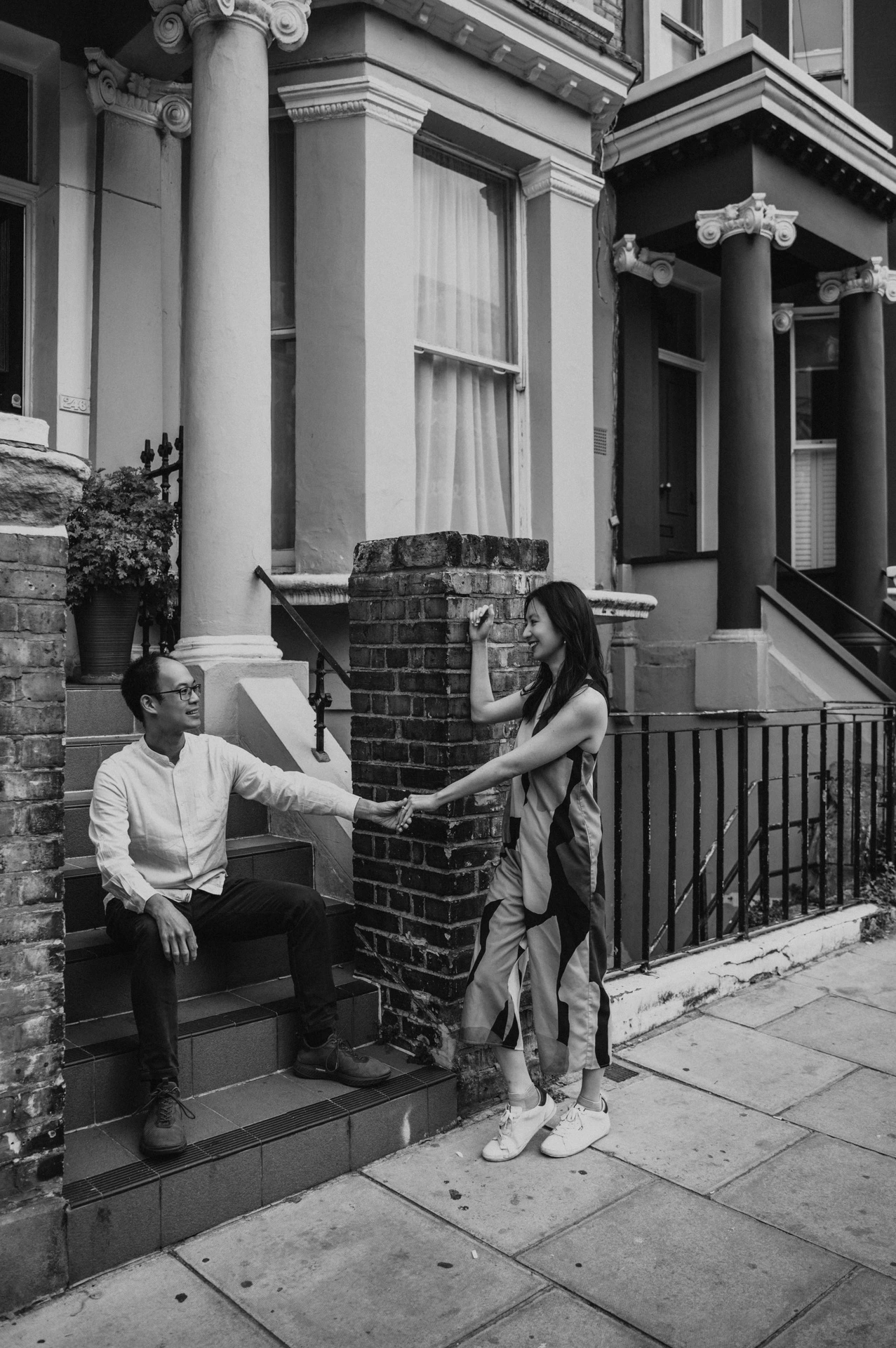 A black-and-white portrait of him seated on the steps holding her hand as she stands beside the brick gatepost during their London couple photoshoot.