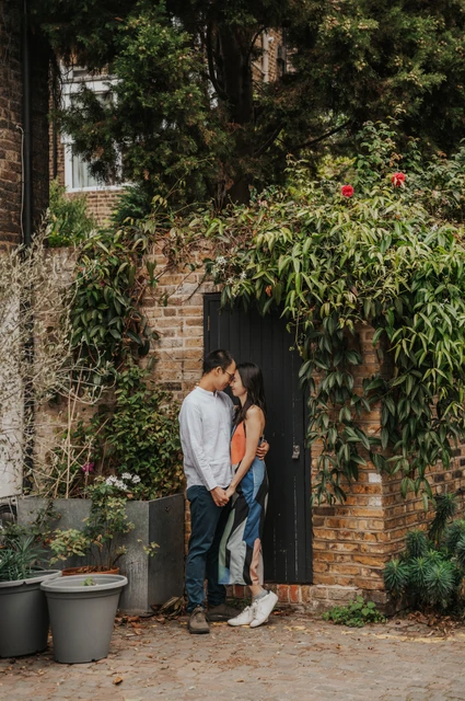 The couple lean close together at a dark door surrounded by climbing greenery, brickwork and plant pots during their London couple photoshoot.