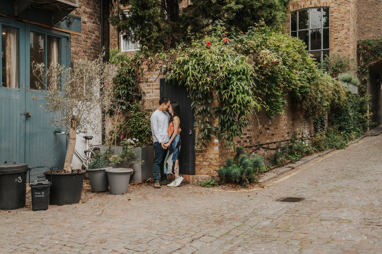 London couple photoshoot: a leafy doorway on a cobbled Notting Hill lane A wide view of the couple standing together at a dark doorway beneath climbing greenery along a cobbled lane during their London couple photoshoot.
