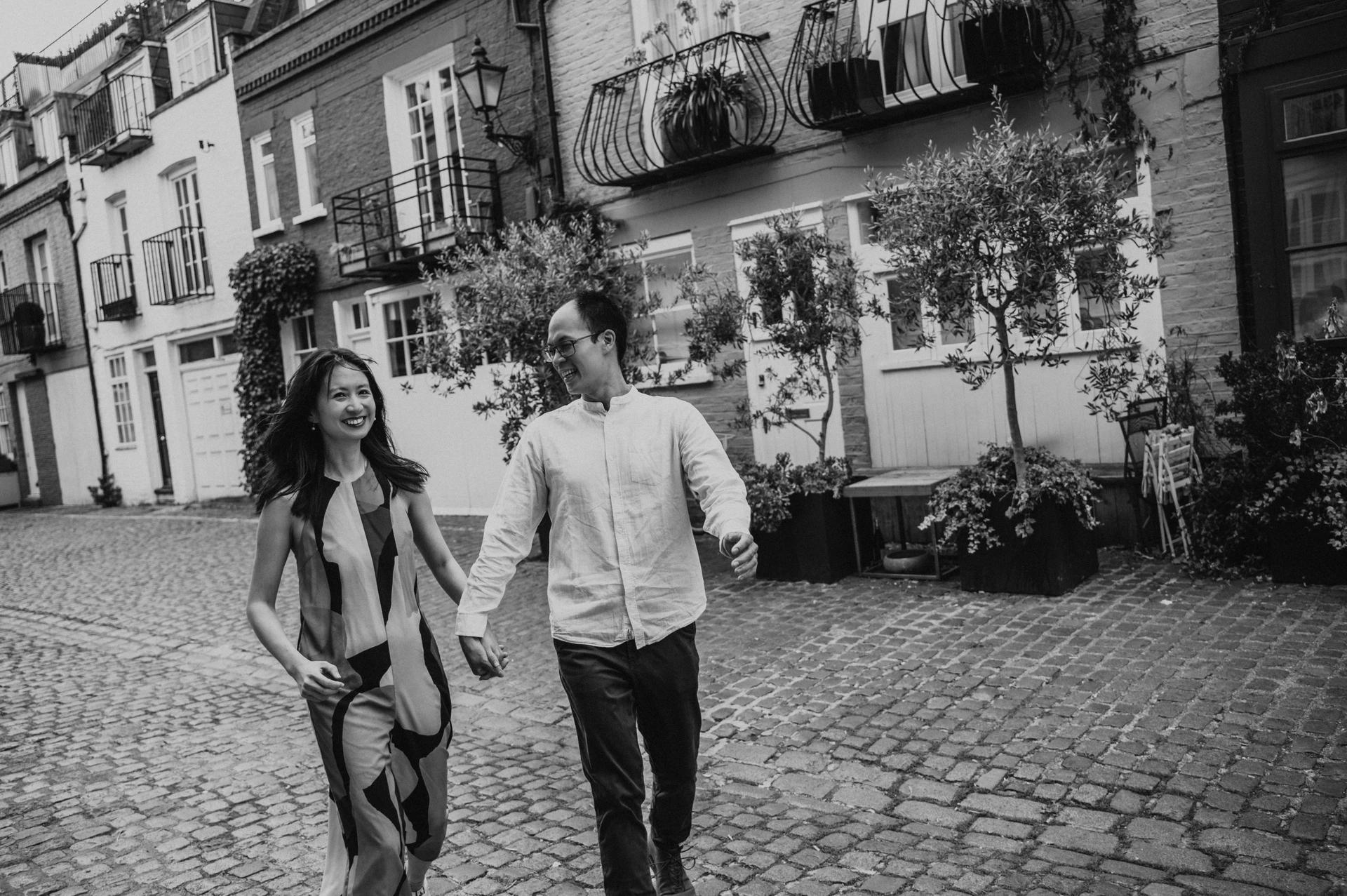 A black-and-white view of the couple smiling at each other as they walk hand in hand along a cobbled Notting Hill mews during their London couple photoshoot.