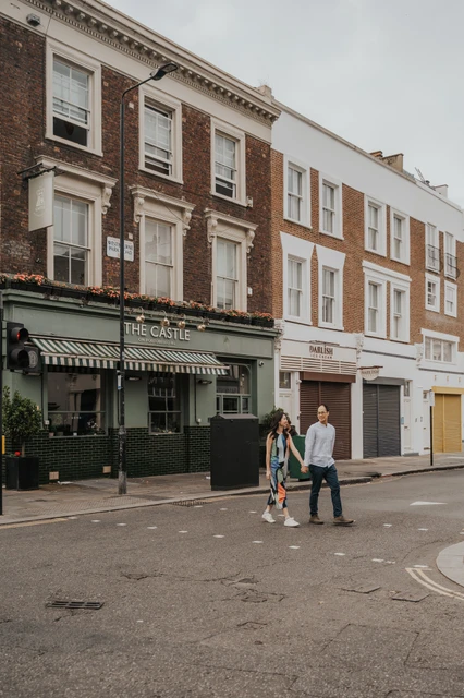 London couple photoshoot: crossing a Notting Hill street together The couple hold hands while crossing a quiet street in front of brick buildings and neighbourhood shopfronts in Notting Hill during their London couple photoshoot.