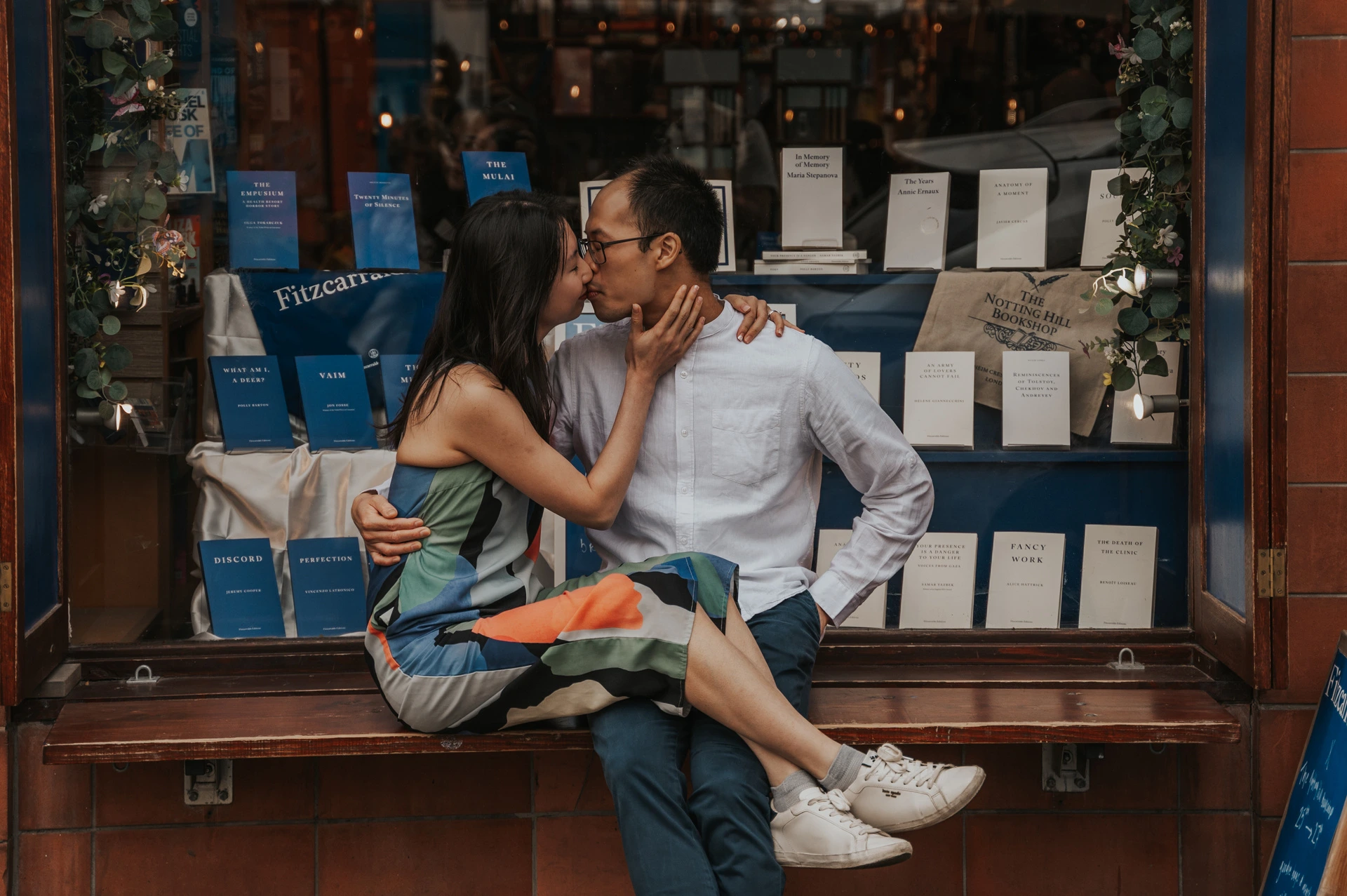 The couple kiss while seated together on the bench in front of the book-filled Notting Hill Bookshop window during their London couple photoshoot.