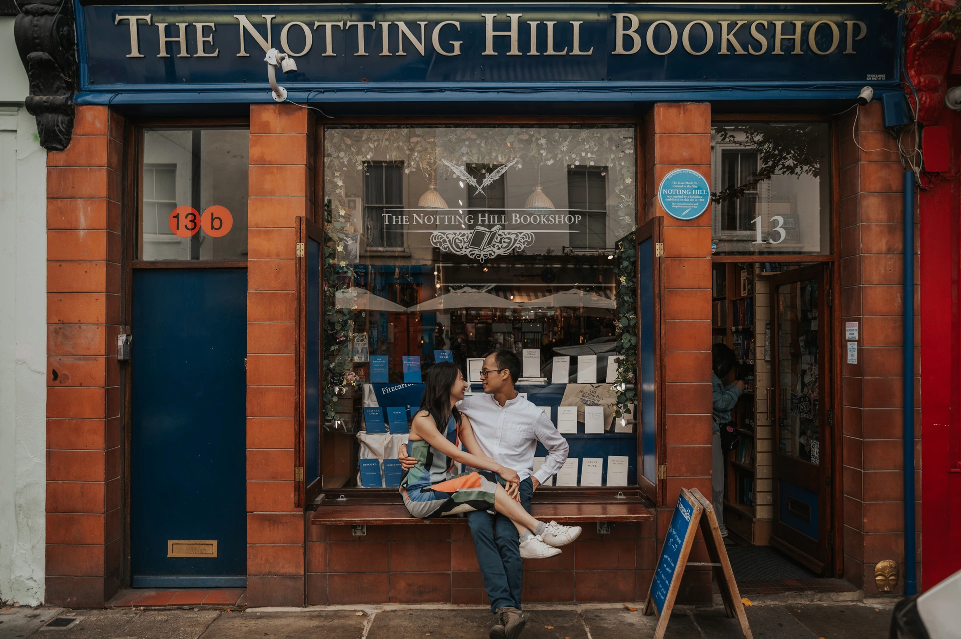 A wide view of the couple seated close together on the bench outside The Notting Hill Bookshop during their London couple photoshoot.
