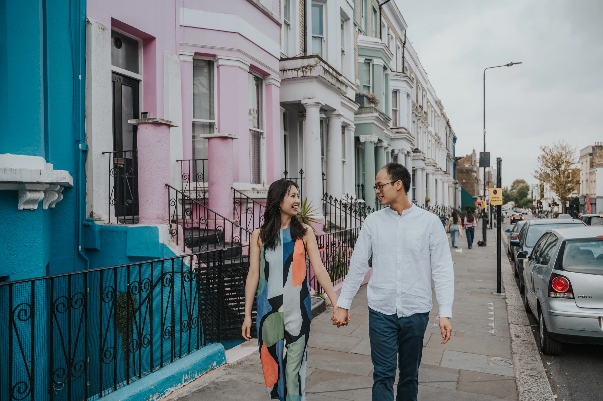 A wide view of the couple walking hand in hand along the pavement beside colourful Notting Hill houses during their London couple photoshoot.