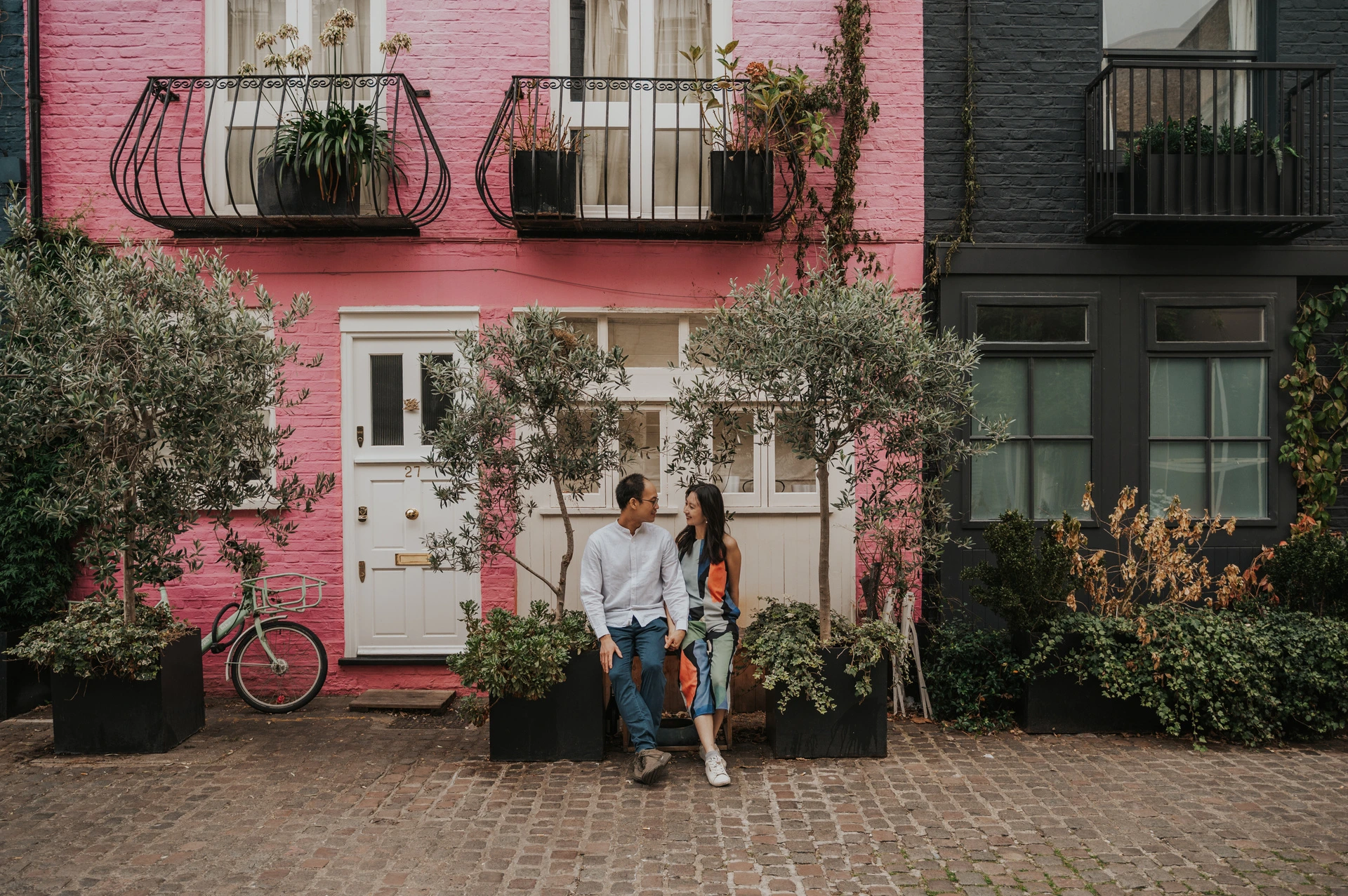 A wide view of the couple sitting outside a pink mews house, framed by potted trees, a bicycle and neighbouring blue paintwork during their London couple photoshoot.