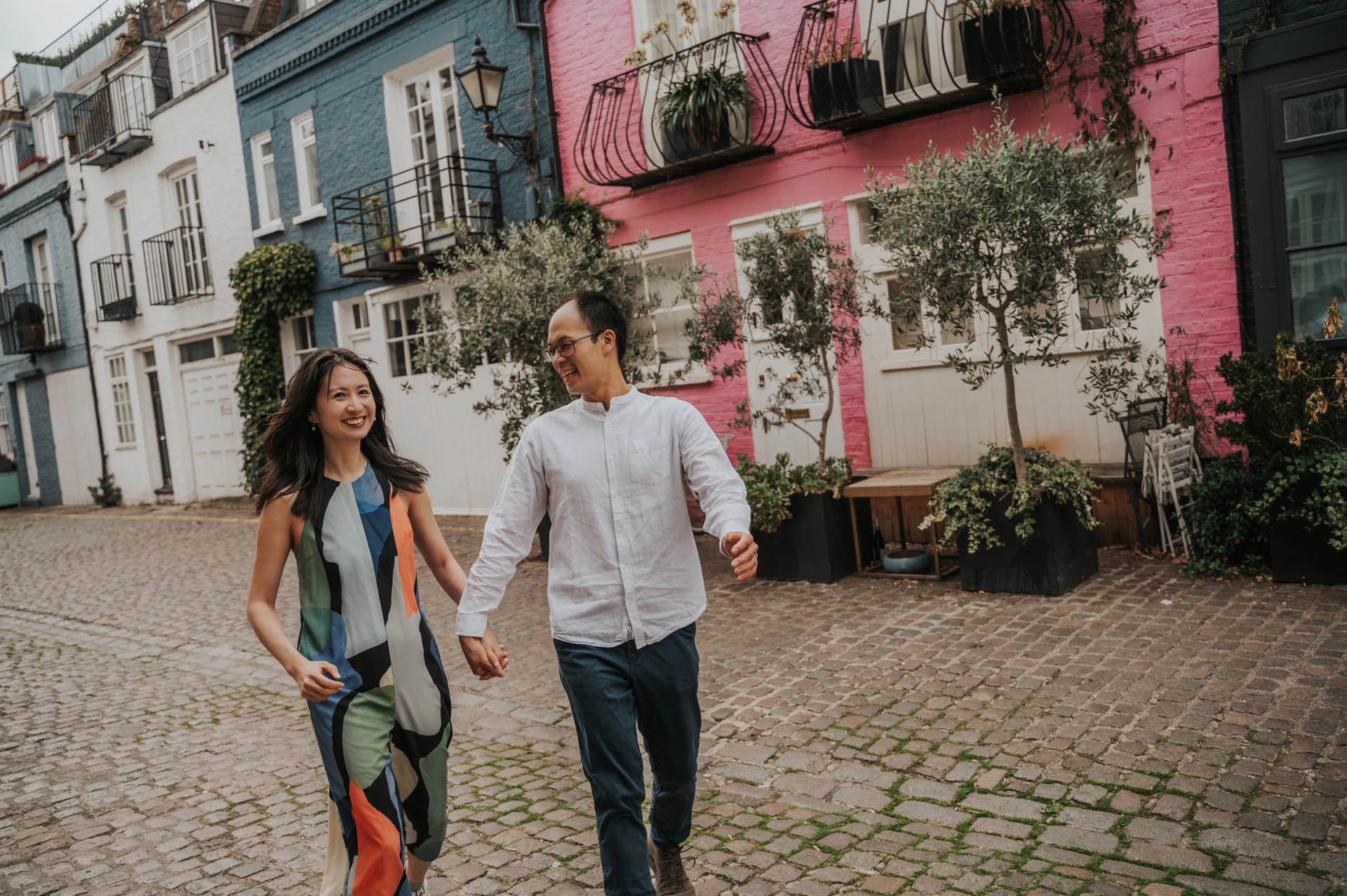 The couple smile at each other while walking hand in hand past pink and blue houses on a cobbled mews during their London couple photoshoot.