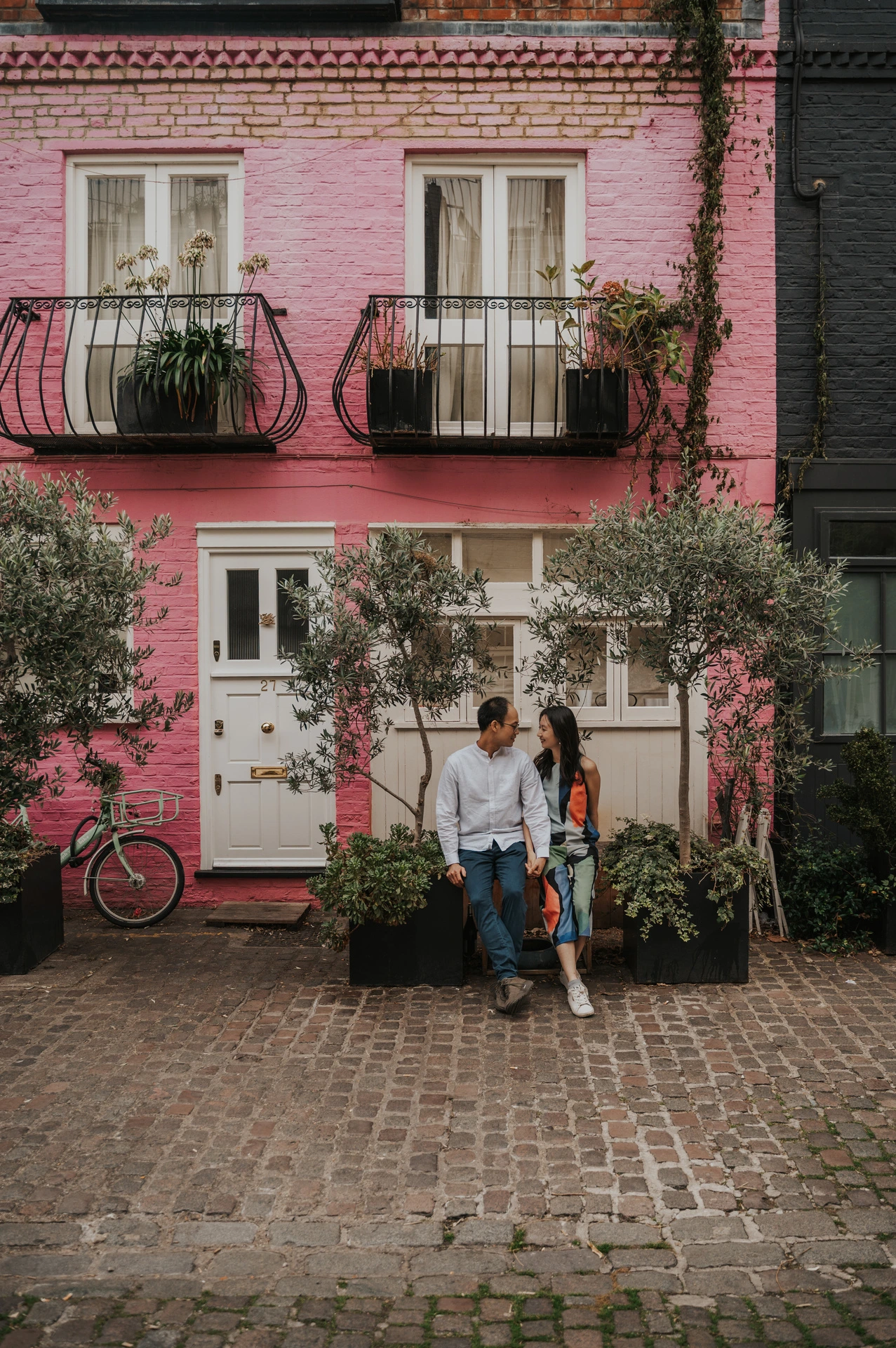 The couple sit close together beneath the windows and balconies of a pink Notting Hill mews house during their London couple photoshoot.