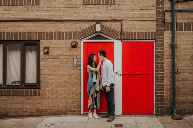 A wide view of the couple standing close together in front of a red garage door set in a brick building during their London couple photoshoot.