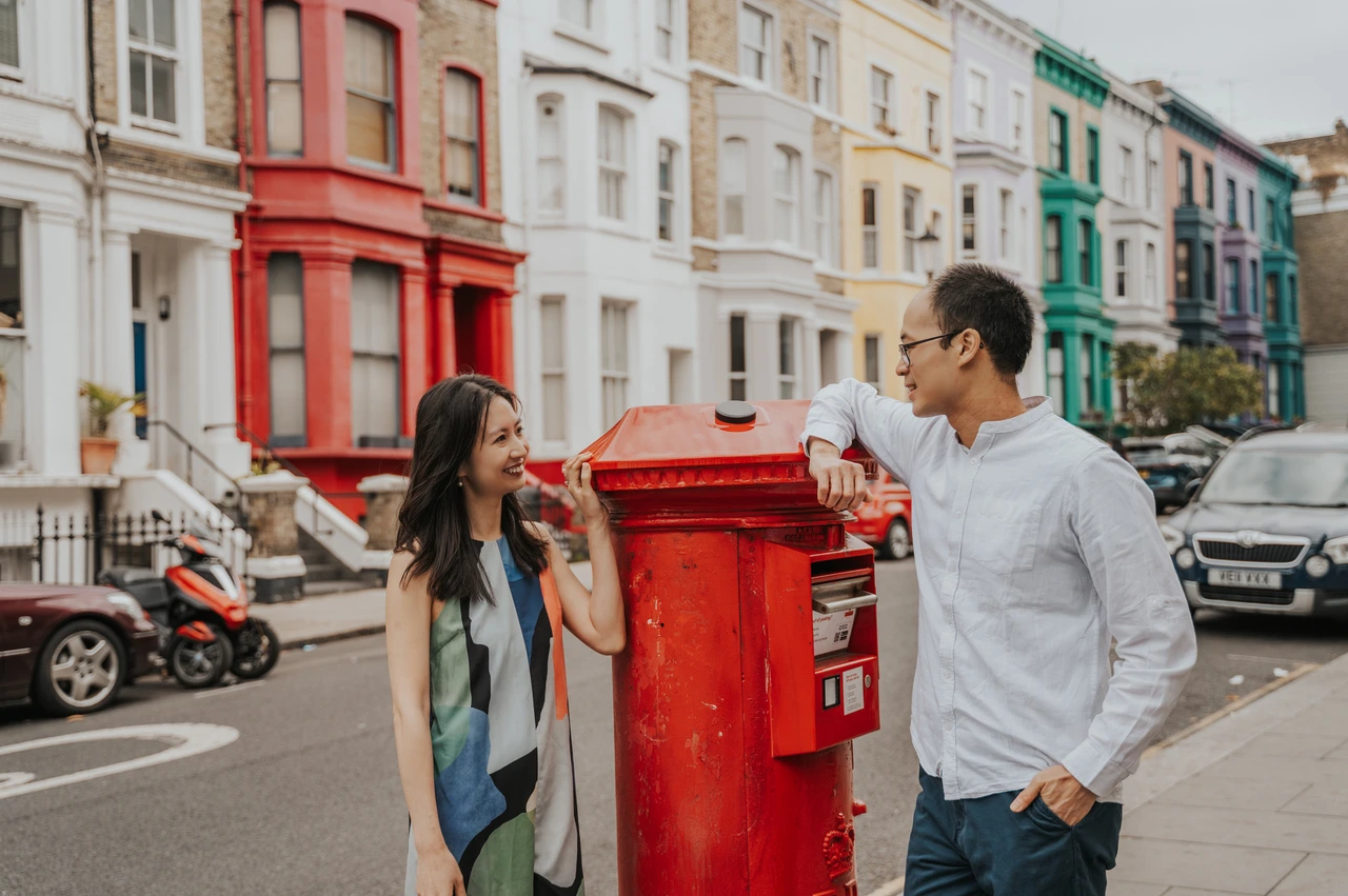 London couple photoshoot: smiling beside a red Notting Hill postbox He rests an elbow on a red postbox while she smiles beside him, with colourful terraced houses behind them during their London couple photoshoot.