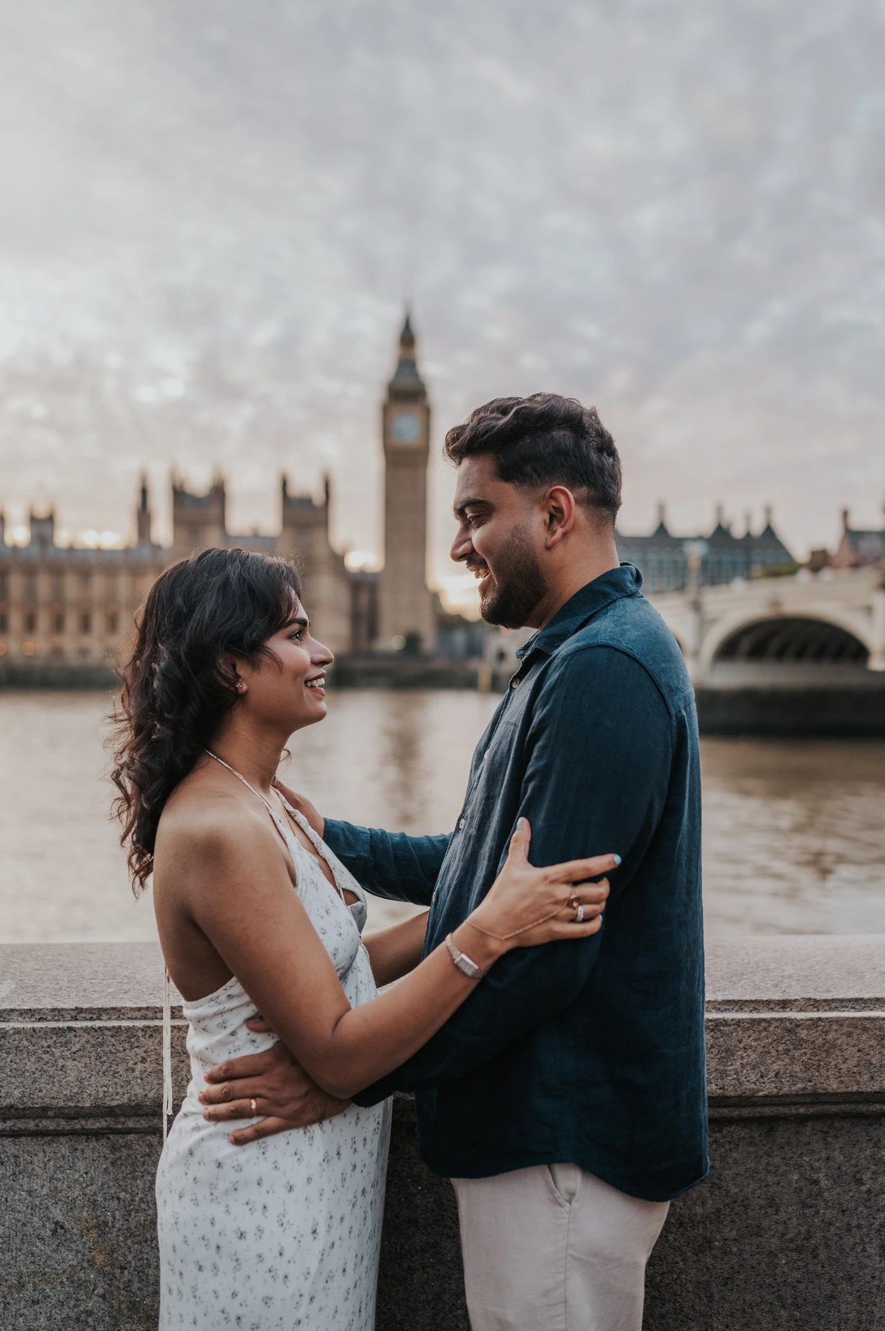 Couple facing one another beside the stone riverside wall, with Big Ben across the Thames, during their London couple photoshoot.