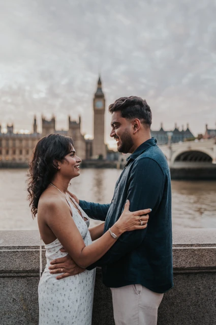 London couple photoshoot face to face by the Thames Couple facing one another beside the stone riverside wall, with Big Ben across the Thames, during their London couple photoshoot.