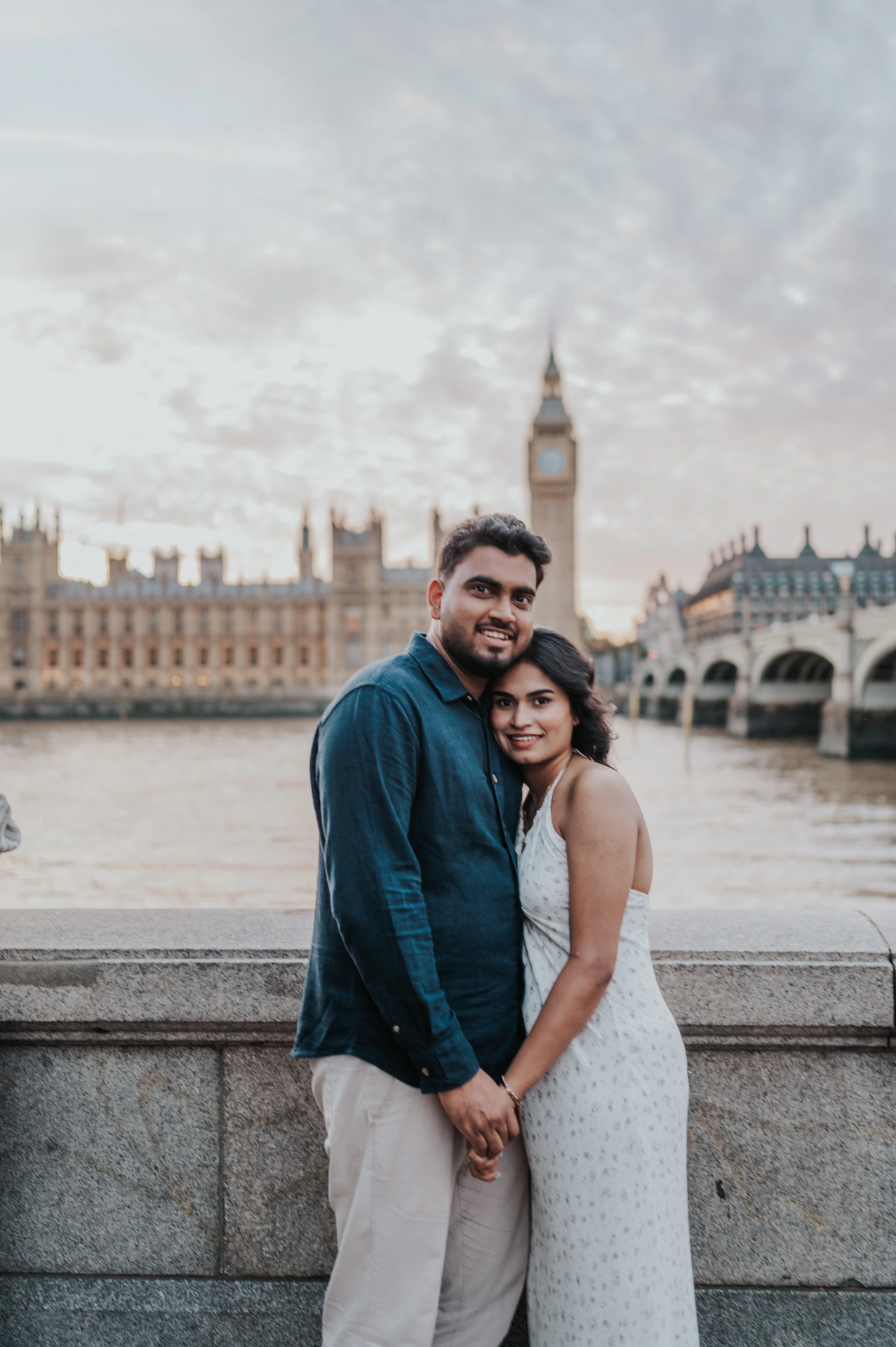 Couple standing close and smiling towards the camera beside the Thames, with Big Ben behind them, during their London couple photoshoot.