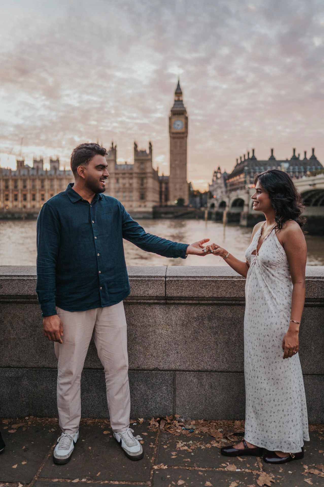 Full-length view of the couple standing apart and holding hands by the riverside wall, with Big Ben behind, during their London couple photoshoot.