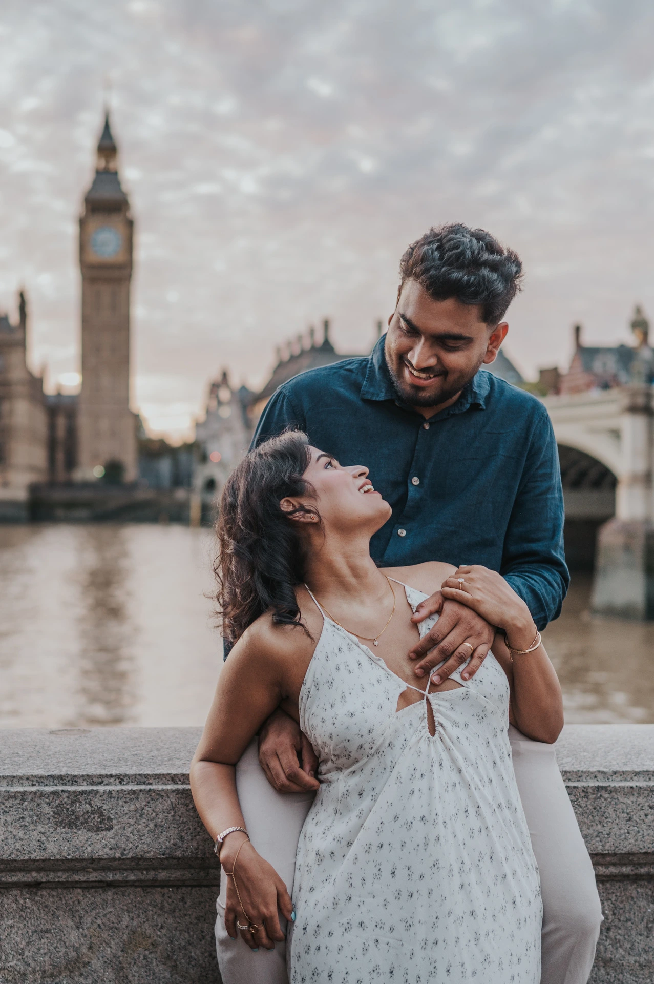 Woman looking up at her partner as he stands behind her beside the Thames, with Big Ben in view, during their London couple photoshoot.
