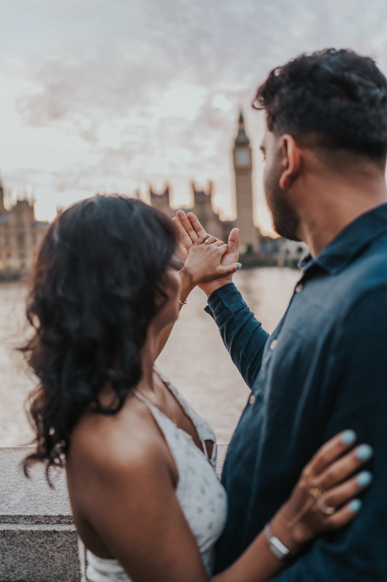 Couple seen over their shoulders looking towards their raised joined hands and Big Ben across the Thames during their London couple photoshoot.