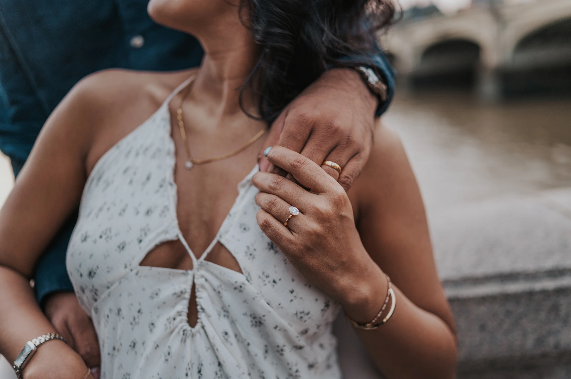 Close view of the couple’s hands at the woman’s shoulder above her pale floral dress, with the river behind, during their London couple photoshoot.