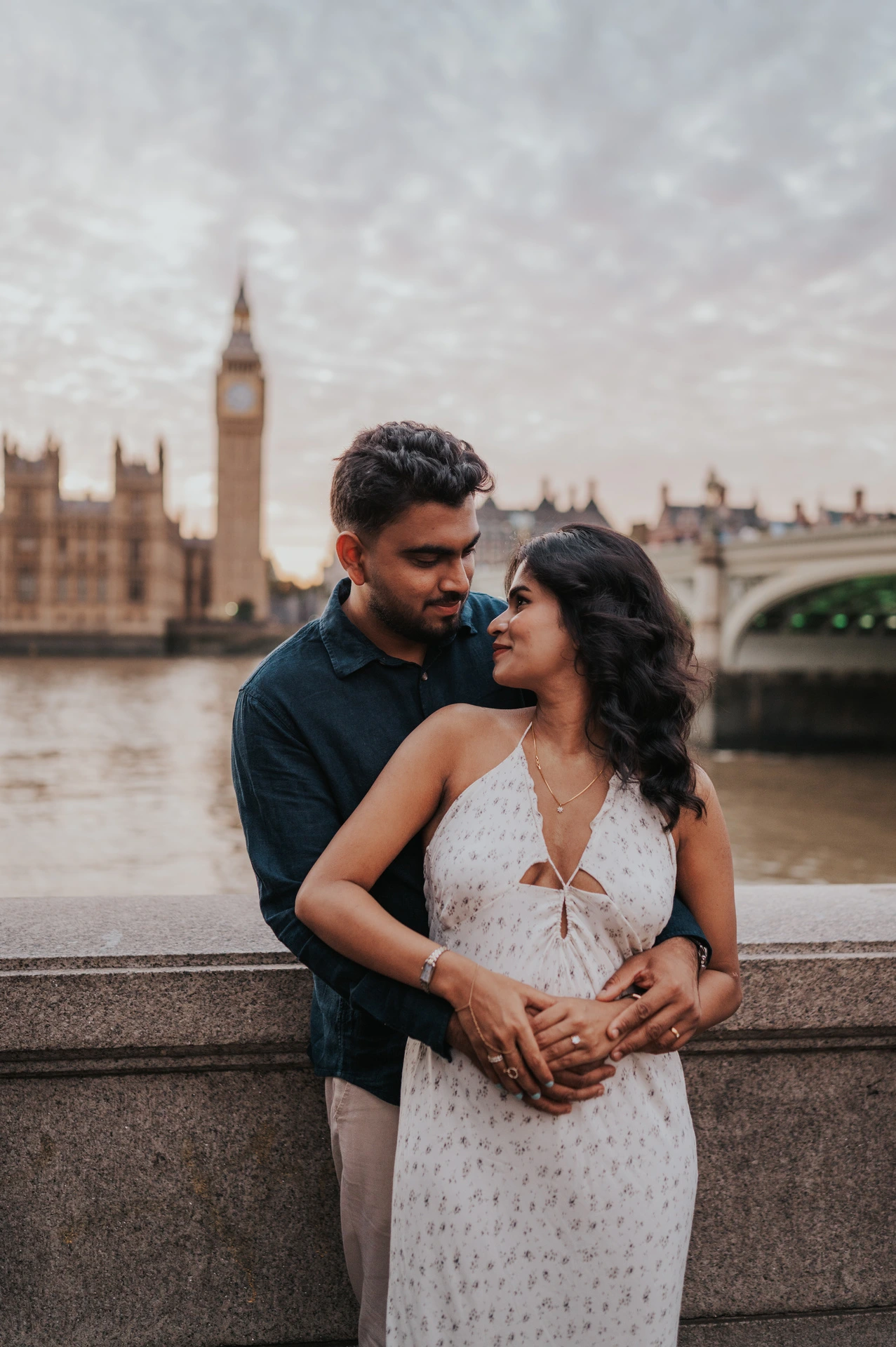 Man standing behind his partner with his arms around her waist as they look at each other by the Thames during their London couple photoshoot.