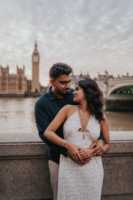 London couple photoshoot in an embrace beside the Thames Man standing behind his partner with his arms around her waist as they look at each other by the Thames during their London couple photoshoot.