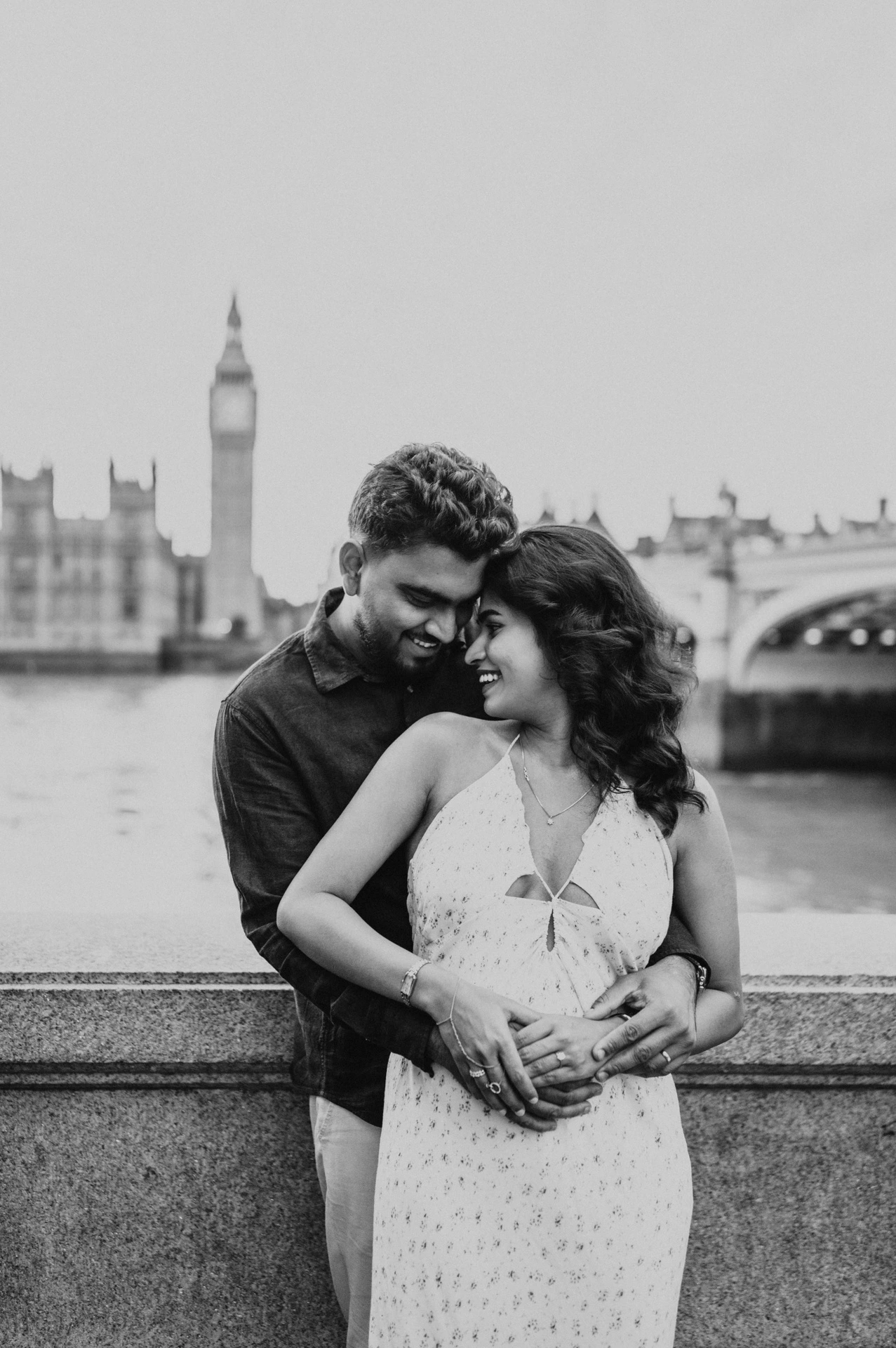 Black-and-white portrait of the couple smiling together in an embrace beside the Thames, with Big Ben behind, from their London couple photoshoot.
