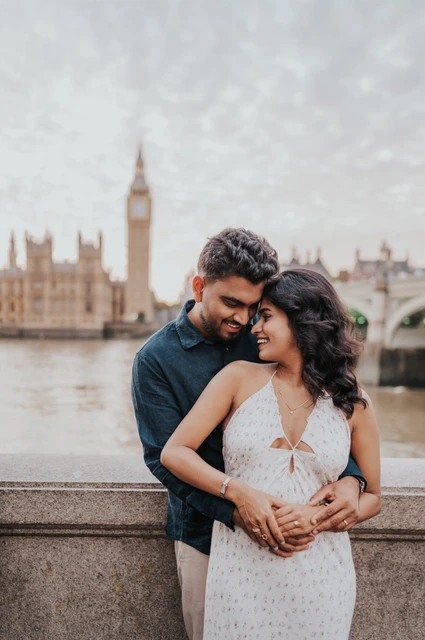 London couple photoshoot smiling together by the river Couple smiling with their heads inclined together and his arms around her waist beside the Thames during their London couple photoshoot.