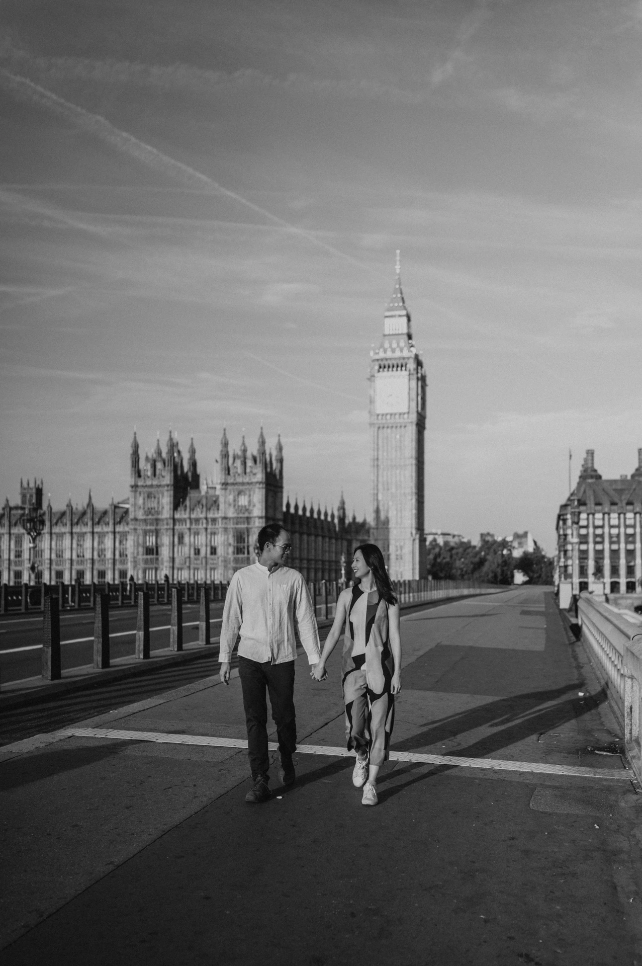 Black-and-white full-length view of the couple walking along Westminster Bridge with Big Ben behind them, from their London couple photoshoot.