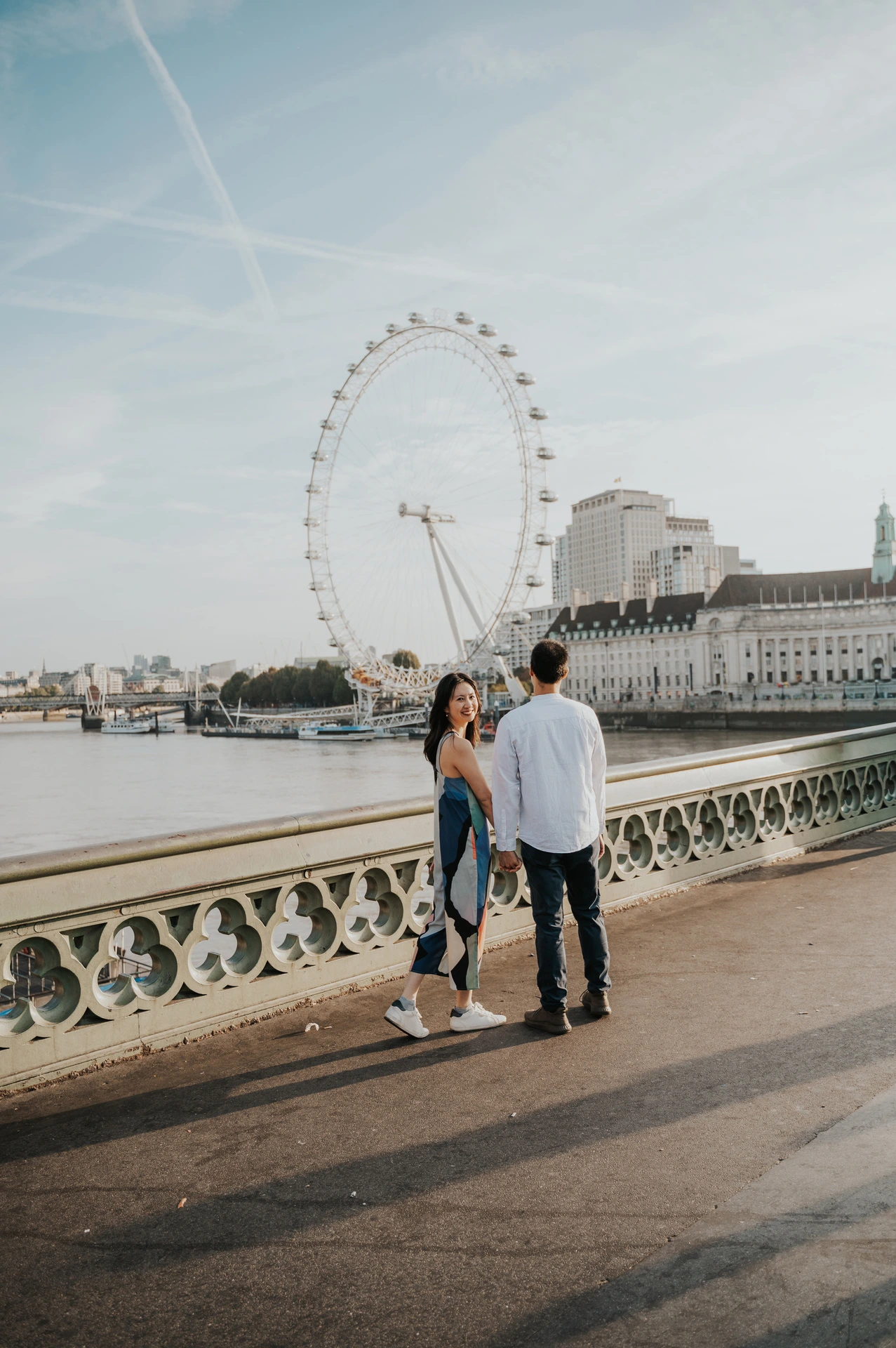 A couple holding hands on Westminster Bridge, with the woman looking back at the camera and the London Eye across the river during their London couple photoshoot.