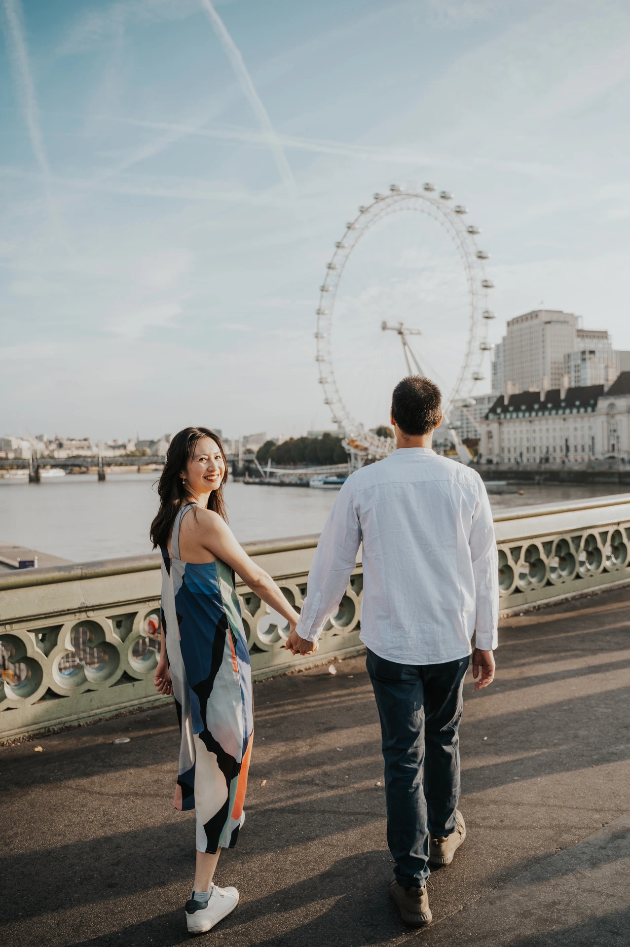 Couple holding hands on Westminster Bridge, with the London Eye above the Thames behind them, during their London couple photoshoot.