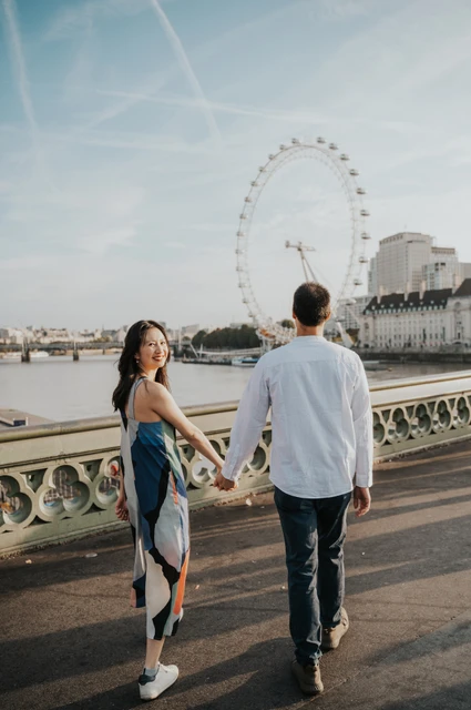 London couple photoshoot walking with the London Eye behind Couple holding hands on Westminster Bridge, with the London Eye above the Thames behind them, during their London couple photoshoot.