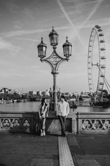 Black-and-white wide view of the couple beneath an ornate Westminster Bridge lamp, with the London Eye in the background, from their London couple photoshoot.
